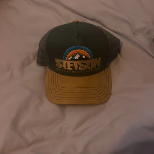 Stetson keps  - Snygg truckerkeps från Stetson med broderat naturmotiv och texten 'Exploring Nature Since 1865'. Framsidan är mörkgrå med detaljer i blått och orange, och skärmen är beige. Baksidan har mesh i grått för extra ventilation. Perfekt för dig som gillar outdoor och cool stil.