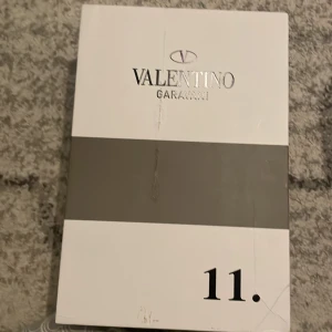 Valentino Garavani Open sneakers vit/svart - Snygga Valentino Garavani Open sneakers i vitt skinn med svart band och silverfärgade nitar. Klassisk rund tå, platt vit sula och diskret logga på plösen. Perfekta för dig som vill ha en lyxig och stilren look med en twist.