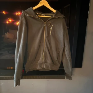 Sail Racing Hoodie - Stilren svart hoodie från Sail Racing med dragkedja framtill och en bröstficka prydd med logga. Den har huva, långa ärmar och är mjukt fleecefodrad på insidan – perfekt för svalare dagar. Kombinera med jeans eller joggers för en avslappnad och trendig look. Inga defekter!