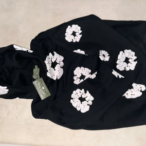 Svart hoodie från Denim Tears med tryck - Svart hoodie från Denim Tears med unika vita grafiska tryck över hela tröjan. Klassisk huva och avslappnad passform, perfekt för dig som vill sticka ut. Tillverkad i mjuk bomull för maximal komfort.