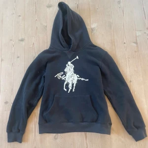 Svart hoodie från Polo Ralph Lauren - Svart hoodie från Polo Ralph Lauren med stor vit logga och polospelare på bröstet. Tröjan har huva, magficka och ribbade muddar vid ärmslut och nederkant. Mjuk och skön bomullsmix, perfekt för chill dagar. Klassisk och stilren design.
