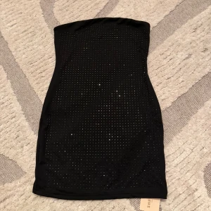 Svart glittrig tubklänning från SHEIN - Snygg svart tubklänning från SHEIN med glittriga detaljer framtill. Klänningen är kort och figurnära, perfekt för dig som vill sticka ut. Gjord i stretchigt syntetmaterial som sitter skönt på kroppen. Enkel och stilren baksida utan detaljer. Oanvänd med lappen kvar på.