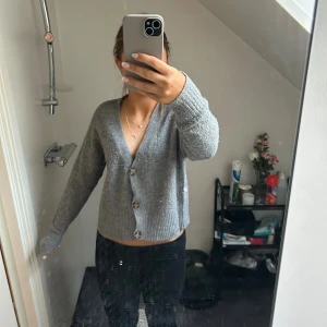 Grå stickad kofta med knappar - Mysig grå stickad kofta med v-ringning och stora knappar framtill. Modellen är lite croppad och har långa ärmar. Perfekt att slänga på sig över ett linne eller t-shirt för en chill look.