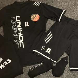 Svart Unihoc Hawks fotbollströja - Säljer träningskläder från Unihoc med vita tryck och detaljer, samt Hawks-logga på ryggen och klubbmärke på bröstet. Tröjan är kortärmad och tillverkad i ett lätt, sportigt material som andas. Perfekt för träning eller match. Träningskläderna, shorts, tröjan och ett par strumpor helt nya. Hoodie och andra paret strumpor i fint skick. Storlek Hoodie 154-160 och resten i storlek 160
