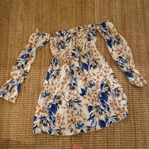 Blommig offshoulder kortklänning - Superfin kortklänning med offshoulder och långa ärmar. Vit botten med blått och orange blommönster. Klänningen har resår i midjan och ärmarna, vilket ger en snygg passform. Perfekt för varma dagar och sommarhäng.