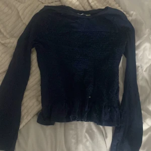 Mörkblå blus med volangkant - Säljer en mörkblå blus med långärmade ärmar och smockad, ribbad mittdel. Blusen har en söt volangkant nedtill och är tillverkad i ett mjukt, lätt material som ger en skön känsla. Perfekt för dig som gillar stilrena detaljer och enkel design. Dock så har den ett slitet mönster där uppe🩷