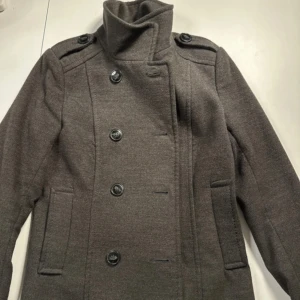 Mörkgrå kappa med hög krage - Snygg mörkgrå kappa med dubbelknäppning och hög krage. Jackan har långa ärmar, axelklaffar och två sidofickor. Perfekt för dig som gillar klassisk stil med en modern twist.