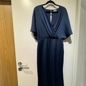 Marinblå långklänning från Bubbelroom - Snygg marinblå långklänning med omlott framtill och v-ringad hals. Klänningen har vida, korta ärmar och en elegant öppning i ryggen med knapp. Materialet är mjukt och har en lätt glansig finish som ger en lyxig känsla.