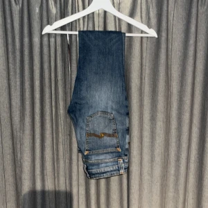 Nudie Jeans blå jeans - Säljer ett par klassiska blå skinny jeans från Nudie Jeans med orange sömmar och slitningar på knäna. Jeansen har fem fickor och är tillverkade i mjukt denimtyg. Perfekta för dig som gillar en stilren look. Byxorna är använda en gång. 10/10 skick. De passar som w 27. Inga frågor är dumma!! 