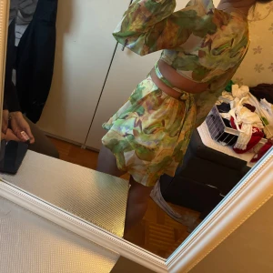 Grön mönstrad omlottklänning med cut out - Superfin kort set med shorts  med grönt och gult abstrakt mönster, djup v-ringning och cut out-detaljer i midjan. Långa, luftiga ärmar och omlottknytning i midjan. Perfekt för dig som vill sticka ut med en färgglad och trendig look.