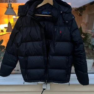 Svart pufferjacka från Polo Ralph Lauren - Svart pufferjacka från Polo Ralph Lauren med huva och flera dragkedjor. Jackan har det klassiska röda Polo-logot på bröstet och är tillverkad i syntetmaterial. Passformen är normal och jackan är perfekt för kalla dagar. Jackan är i väldigt gott skick och har har tagits handom under tiden jag haft den!!!