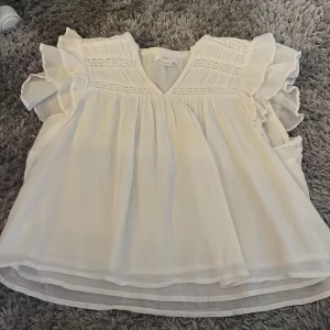 Vit blus med volang från Gina Tricot - Supersöt vit blus från Gina Tricot i storlek 146/152. Blusen har v-ringning, volangdetaljer på axlarna och fina broderade partier upptill. Den är ärmlös och har en luftig, loose passform som ger en chill och fräsch vibe. Perfekt för vår och sommar!