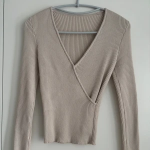 Beige ribbad omlottopp med v-ringning - Beige ribbad omlottopp. I väldigt tjockt och mjukt material perfekt till höst / vintern. Vet inte märket då jag köpte den second hand utomlands i det oanvända sortimentet. Används bara 2 gånger, så är som ny. Skriv om du har frågor💗