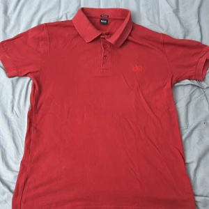 Röd pikétröja från Hugo Boss - Snygg röd pikétröja från Hugo Boss med klassisk krage och tre knappar framtill. Tröjan har korta ärmar och broderad logga på bröstet. Tillverkad i mjuk pima bomull som känns skön mot huden. Perfekt för dig som gillar stilrena och tidlösa plagg.