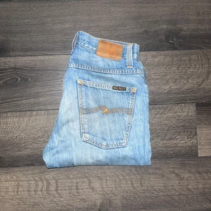 Nudie Grim Tim jeans  - Säljer ett par ljusblå jeans från Nudie Jeans med klassisk femficksdesign. Modell GRIM TIM.  Midja 38cm, längd 103cm. OBS se minimala hål vid bakfickorna på sista bilden. Ifall det inte finns någon bild i annonsen där jag bär plagget så är det för att jag ej kan ha storleken heller passformen, så kolla måtten för storlek och googla på modellnamnet för passform 😉S3