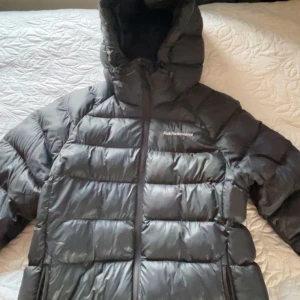 Svart dunjacka Peak Performance - Svart dunjacka från Peak Performance med huva och två dragkedjeförsedda fickor. Jackan har en quiltad design med breda paneler och en snygg, sportig look. Perfekt för kalla dagar och har en normal passform. Logga på bröstet.