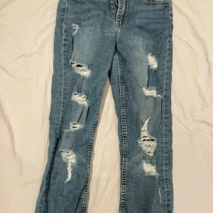 Blå slitna jeans med raka ben - Säljer ett par blå jeans med flera slitningar och hål framtill för en cool, avslappnad look. Jeansen har raka ben och klassiska bakfickor. Perfekta för dig som gillar streetstyle och vill ha ett par jeans med attityd.