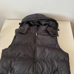 Svart dunväst med huva - Svart dunväst med quiltad design och avtagbar huva. Västen har hel dragkedja framtill och två fickor med dragkedja. Perfekt för lager-på-lager och håller dig varm under kyliga dagar. Helt ny.