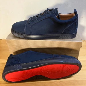 Mörkblå mocka sneakers från Louboutin - Snygga mörkblå sneakers i mocka från Christian Louboutin med klassisk röd sula och broderad logga på hälen. Skorna har rund tå, snörning och en stilren design som sticker ut med den ikoniska röda undersidan.