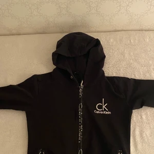 Svart hoodie från Calvin Klein Jeans - Svart hoodie från Calvin Klein Jeans med vit ck-logga på bröstet och dragkedja framtill. Tröjan har huva och långa ärmar, samt snörning vid midjan. Perfekt för dig som gillar stilrena och klassiska streetwear-plagg.