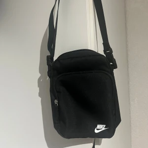Svart axelväska från Nike - En stilren svart axelväska från Nike med justerbar rem och klassisk vit logga framtill. Väskan har ett huvudfack och en mindre ficka med dragkedja på framsidan, perfekt för mobilen och andra småprylar. Tillverkad i slitstark polyester.