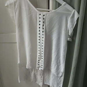 Vit blus med nitar och kort ärm - Snygg vit blus med korta ärmar och dekorativa silverfärgade nitar längs en lodrät söm framtill. Blusen har rund halsringning och är tillverkad i ett tunt, mjukt tyg som faller fint. Perfekt för en trendig och avslappnad look.