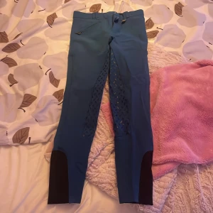 Blå ridbyxor med silikon och svarta detaljer - Snygga blå ridbyxor med silikonmönster på insidan av benen för extra grepp. Byxorna har svarta förstärkningar vid bensluten och en dragkedjeficka framtill. Lapp borta, men vet strl. 1 knapp på framsidan, Säljer då dom är för små för mig.