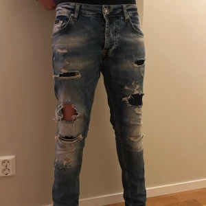 Blå slitna jeans med broderad bakficka. K0PIA MEN VÄLDIGT BRA KVALITET - Säljer ett par blå jeans med kraftiga slitningar och hål på benen. Jeansen har en broderad tiger på ena bakfickan och klassisk femficksdesign. De är i en smal passform och har snygga slitdetaljer för en trendig look. Jag är 184 cm 