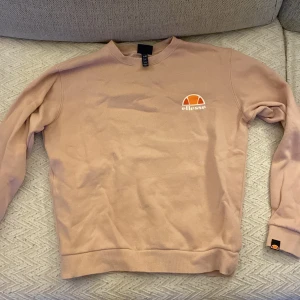 Rosa sweatshirt från Ellesse - Säljer en rosa sweatshirt från Ellesse med klassisk logga i orange och vitt på bröstet och ärmslutet. Tröjan har rund hals, långa ärmar och ribbade muddar. Perfekt för chill dagar och enkel att matcha med jeans eller joggers.