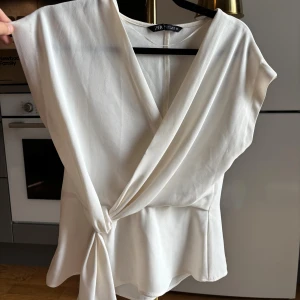 Vit omlottopp med knyt från Zara - Säljer en stilren vit omlottopp från Zara i storlek S. Toppen har v-ringning, knyt i sidan och är ärmlös. Tillverkad i ett lätt och mjukt material som ger en snygg drapering. Perfekt för dig som gillar en clean och chic look.