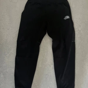 Svarta mjukisbyxor från The North Face - Svarta mjukisbyxor från The North Face med logga på benet och längs sidan. Byxorna har elastisk midja och ribbade muddar vid bensluten. Tillverkade i mjukt material, perfekta för chill eller träning. Den är i storlek S men skulle säga att den inte är normal i storleken den är en XS skulle passa dig som är 160-168 cm