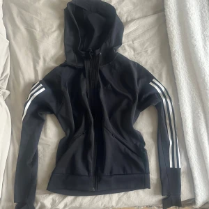Svart Adidas hoodie med dragkedja i XS - Svart hoodie i väldigt fint skick från Adidas i modellen Climalite med dragkedja och huva. Klassiska vita ränder längs ärmarna och Adidas-logga på bröstet. Tillverkad i polyester, perfekt för träning eller chill. Snygg och sportig passform.