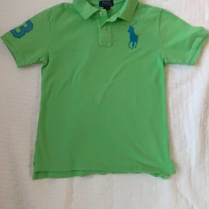Grön pikétröja från Ralph Lauren - ungdom - Snygg grön pikétröja från Ralph Lauren med klassisk krage, knappar och blå logga på bröstet. På ena ärmen finns en stor blå siffra 3. Tröjan är i bomull och har korta ärmar.
