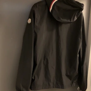 Svart windbreaker från Moncler - Storlek 3 passar m och s. Svart vindjacka från Moncler med huva som har röd och vit rand. Jackan har dragkedja framtill, lång ärm och Moncler-logga på vänster arm. Materialet är lätt och syntetiskt, perfekt för blåsiga dagar. Snygg och stilren design med normal passform.