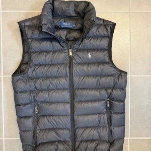 Svart dunväst från Polo Ralph Lauren - Snygg svart dunväst från Polo Ralph Lauren med horisontella quiltade paneler och klassisk logga broderad på bröstet. Västen har hög krage, två fickor med dragkedja och hel dragkedja framtill. Perfekt för lager-på-lager och chill stil.