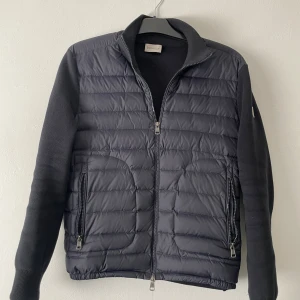 Svart quiltad pufferjacka Moncler - Svart pufferjacka från Moncler med quiltad framsida i syntet och stickade ärmar. Jackan har två sidofickor med dragkedja, ribbad krage och dubbla dragkedjor framtill. Moncler-logga på vänster ärm ger en clean och exklusiv vibe.