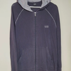 Mörkblå hoodie från Hugo Boss - Snygg mörkblå hoodie från Hugo Boss med ljusgrå huva och vita detaljer längs axlarna. Tröjan har dragkedja framtill, två fickor och diskret Boss-logga på bröstet. Perfekt för chill dagar och streetwear-stil. Ganska urtvättad men ser mer ut på bild. I fint skick annars!