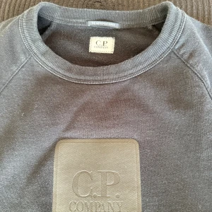 Svart sweatshirt från C.P. Company - Svart sweatshirt från C.P. Company med rund hals och ett stort, stilrent logopatch på bröstet. Tröjan har långärmade raglanärmar och ribbade muddar vid hals, ärmslut och nederkant. Perfekt för dig som gillar clean streetwear och snygga detaljer.