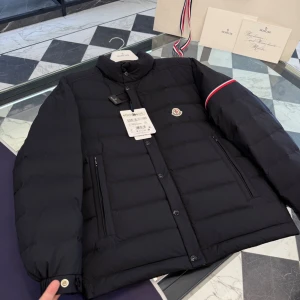 Svart Moncler dunjacka herr - Svart dunjacka från Moncler med quiltad design, ståkrage och ikonisk logga på bröstet. Jackan har dragkedja och knappar framtill, samt två sidofickor med dragkedja. Röd, vit och blå detalj på ena ärmen ger extra stilpoäng. Perfekt för kalla dagar.