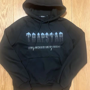Svart Trapstar hoodie med tryck - Svart hoodie från Trapstar med stort silvrigt logotryck på bröstet och en unik detalj med dragkedja över texten. Huva med snörning och tryck även på huvan. Mjuk insida och klassisk känguruficka framtill. Perfekt för dig som gillar streetwear.