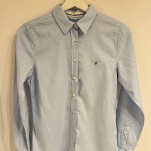 Ljusblå skjorta från Gant, strl 36 - Klassisk ljusblå skjorta från Gant med broderad logga på bröstet. Skjortan har lång ärm, normal passform och knäppning framtill med vita knappar. Perfekt krage och stilren look, tillverkad i mjuk bomull.