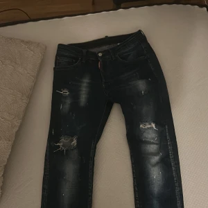 Dsquared2 blå slitna jeans strl 48 - Säljer ett par blå jeans från Dsquared2 i storlek 48. Jeansen har coola slitningar och trasiga detaljer på benen, klassisk femficksmodell och snyggt Dsquared2-märke bak. De är tillverkade i jeansmaterial och har en modern, smal passform.
