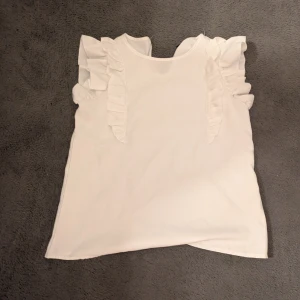 Vit blus med volangärmar - Säljer en söt vit blus med volangdetaljer på axlarna och ärmarna. Blusen har rund hals och är tillverkad i ett mjukt, lätt material som ger en skön känsla. 