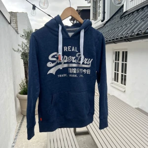Marinblå Superdry hoodie med tryck - Säljer en marinblå hoodie från Superdry med stort vitt tryck på bröstet och japanska tecken på ärmen. Tröjan har huva med vita snören, känguruficka och ribbade muddar. Perfekt för dig som gillar streetwear och snygga detaljer.