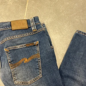 Blå jeans från Nudie Jeans Co - Snygga blå jeans från Nudie Jeans Co med klassisk femficksdesign och kontrastsömmar. Jeansen har en rak passform och är tillverkade i mjukt denimtyg med subtila slitningar. Bakfickan har Nudies ikoniska broderi i orange. W30 l30 9/10 skick 