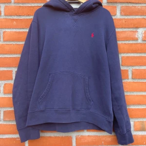 Polo Ralph Lauren Hoodie - Mörkblå hoodie från Polo Ralph Lauren i juniorstorlek XL (18–20), vilket motsvarar ungefär S/M i herrstorlek. Klassisk modell med känguruficka och röd broderad Polo-logotyp på bröstet. Tillverkad i mjuk och bekväm bomullsblandning med borstad insida som gör den varm och skön att bära. Plagget är i mycket fint skick, utan större slitage eller defekter.  Detaljer: 	•	Färg: Mörkblå 	•	Storlek: Junior XL (18–20) ≈ Herr S/M 	•	Mått: Axelbredd 45 cm, längd 60 cm 	•	Märke: Polo Ralph Lauren 	•	Broderad