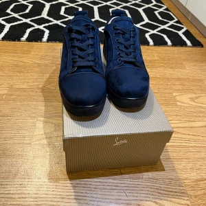 Mörkblå sneakers från Christian Louboutin - Snygga mörkblå sneakers från Christian Louboutin i mocka med klassisk röd sula och diskret logga. Skorna har rund tå, snörning och en stilren design som sticker ut med den ikoniska röda undersidan. Perfekta för dig som vill ha något exklusivt och trendigt.