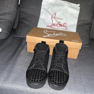 Christian Louboutin svarta sneakers med nitar - Svarta sneakers från Christian Louboutin med mattsvarta nitar på tån, klassisk snörning och ikonisk röd sula. Skorna är tillverkade i mocka och har en rund tå. Perfekta för dig som vill sticka ut med en edgy och lyxig stil.