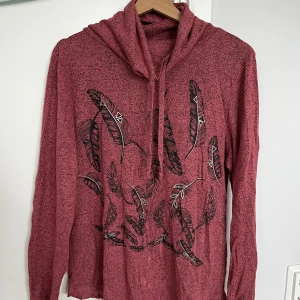 Rosa hoodie med fjäderprint och snörning - Säljer en chill rosa hoodie med svart fjädermönster framtill. Tröjan har huva med snörning och långa ärmar. Materialet är mjukt och känns som bomull eller polyester. Perfekt för dig som gillar lite boho-vibe och vill ha något bekvämt och snyggt.