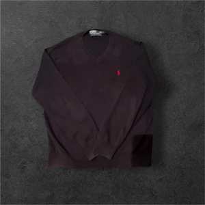 Svart v-ringad tröja från Ralph Lauren - Svart v-ringad tröja från Ralph Lauren med klassisk röd broderad logga på bröstet. Tröjan har lång ärm och är tillverkad i mjuk bomull, perfekt för lager-på-lager. Enkel och stilren design som funkar till det mesta.